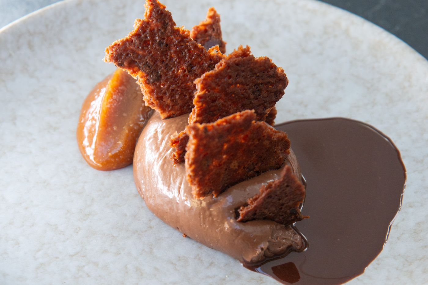 Dessert au chocolat : quenelle de mousse au chocolat, caramel et éclats de biscuit, nappé de sauce chocolat dans une assiette en grès mouchetée
