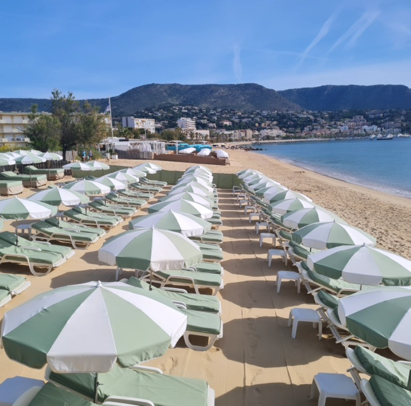 La plage privée de L'Effet Mer : rangées de transats et parasols vert d'eau sur le sable, face à la baie du Lavandou