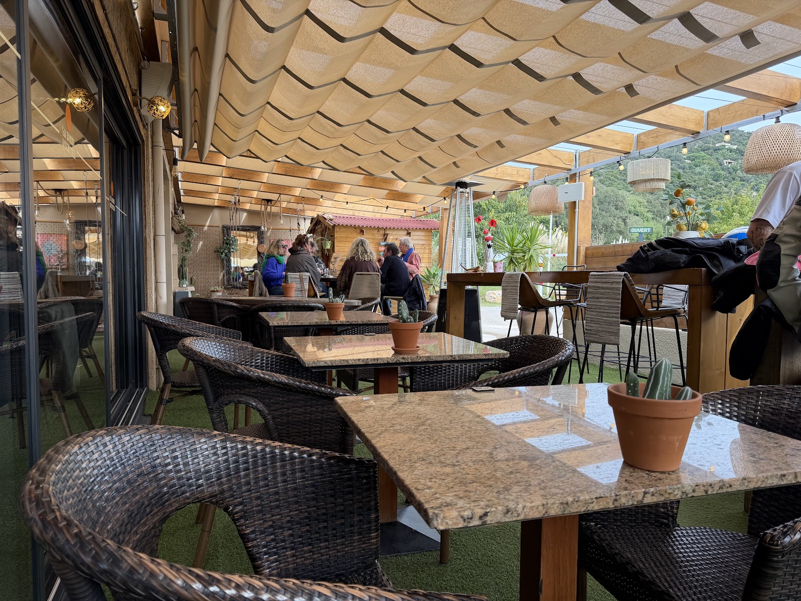 La terrasse ombragée de La Bonne Aventure : tables en marbre, fauteuils en rotin sous la pergola de bois et guirlandes, face à la verdure de Cavalière