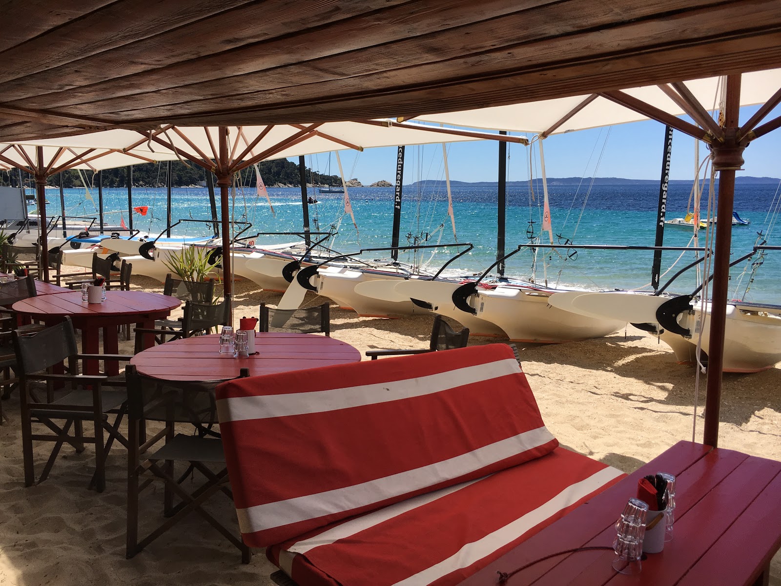 La terrasse de La Cabane sur le sable de Pramousquier : banquettes rayées rouge et blanc sous la pergola, catamarans alignés sur la plage et mer turquoise