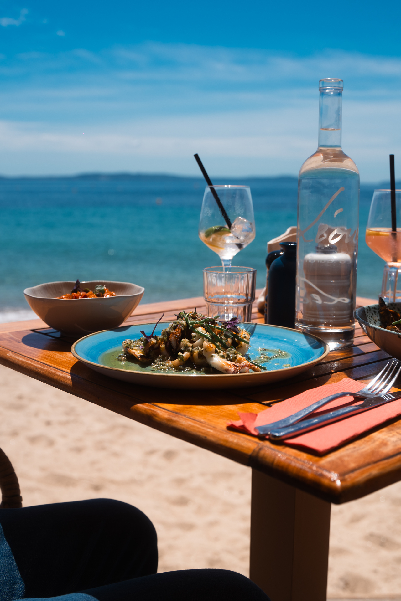 Une assiette généreuse et un verre de rosé sur une table de Noca Plage, sable clair et mer bleue en arrière-plan