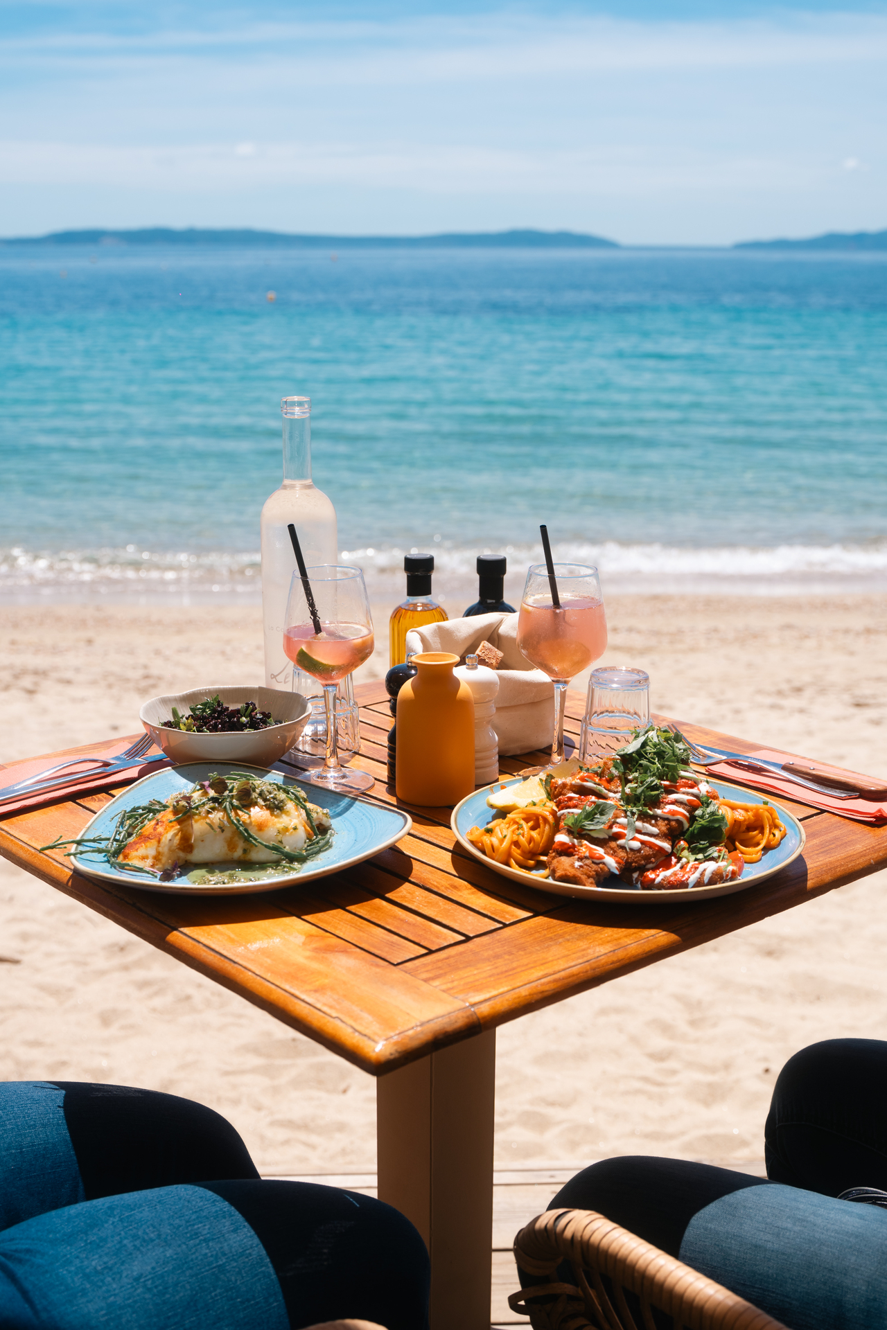 Deux assiettes de poisson et de pâtes accompagnées de rosé, sur une table face au sable et à la mer de Cavalière