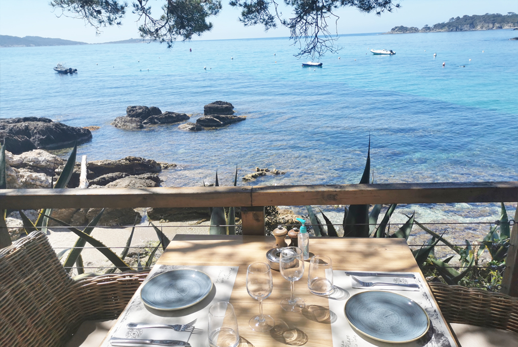 Table dressée sur la terrasse en bois de Pradeau Plage, surplombant une eau turquoise cristalline et de petits bateaux
