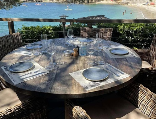 Table ronde en bois dressée avec assiettes en grès bleu et fauteuils en rotin sur la terrasse de Pradeau Plage, face à la crique turquoise et la plage